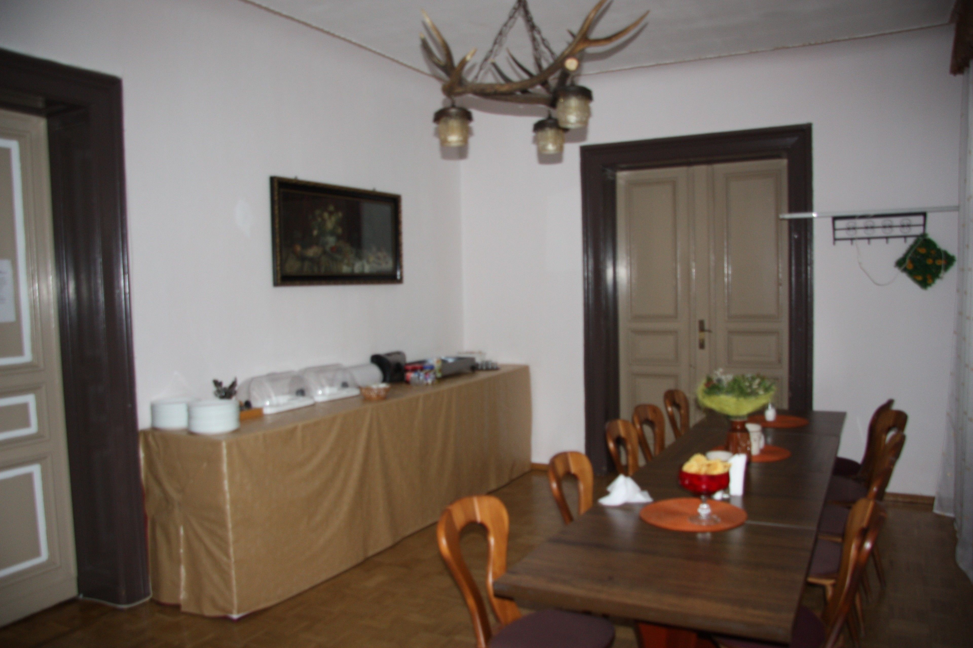 A dining area with a long wooden table, chairs, a buffet table with food items, a chandelier, and a framed picture on the wall.