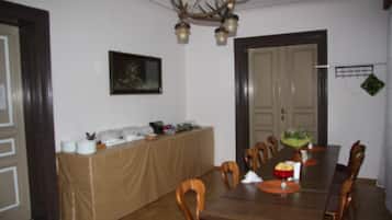 A dining area with a long wooden table, chairs, a buffet table with food items, a chandelier, and a framed picture on the wall.