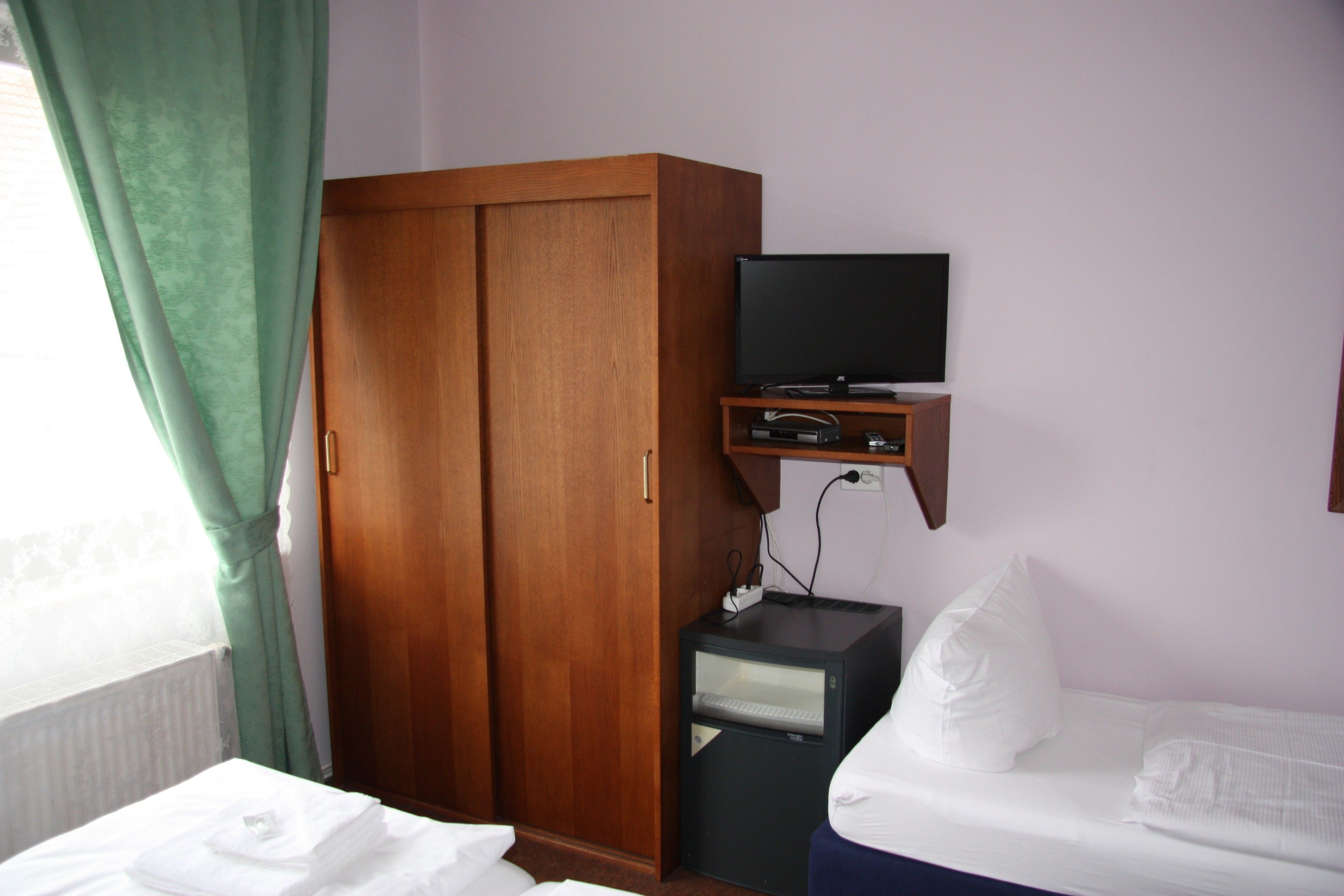 A hotel room with a wooden wardrobe, a television on a stand, a bed with white bedding, and a window with green curtains.