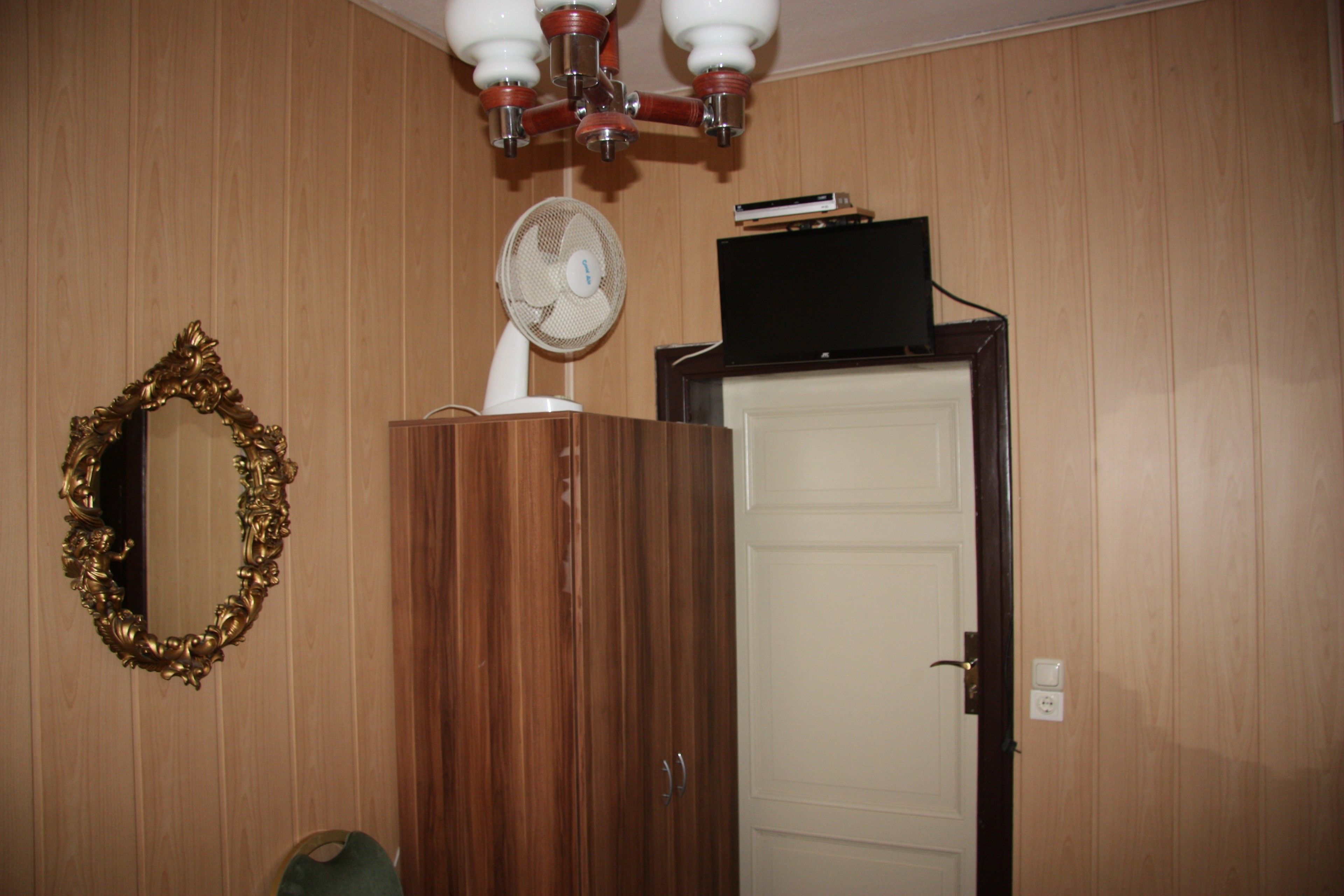 Een kamer met houten paneelwanden, een witte deur, een houten garderobe, een spiegel met een versierde lijst, een plafondventilator en een televisie bovenop de garderobe.