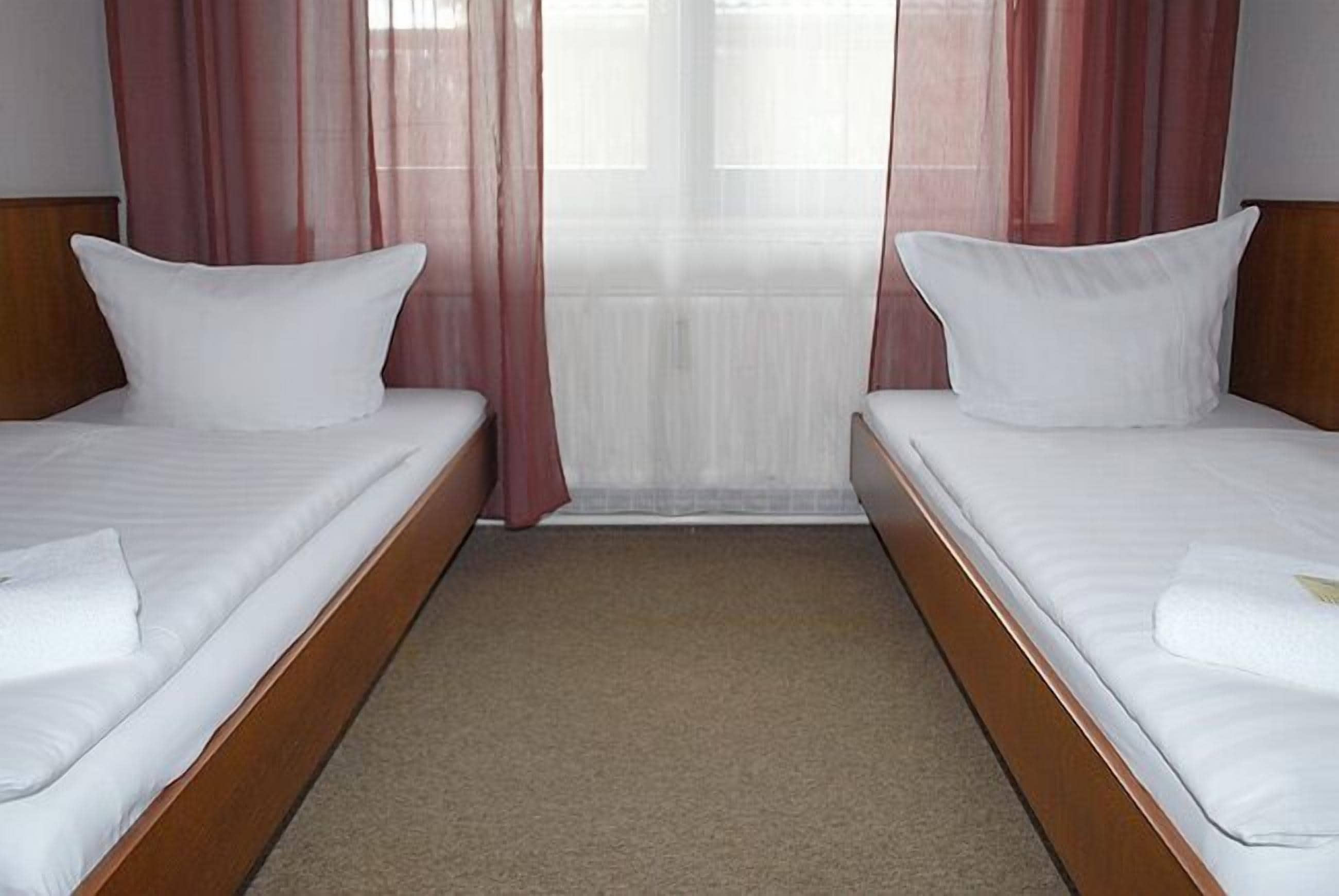 Camera d'albergo con due letti singoli, una testiera in legno, pavimento tappezzato e tende leggere.