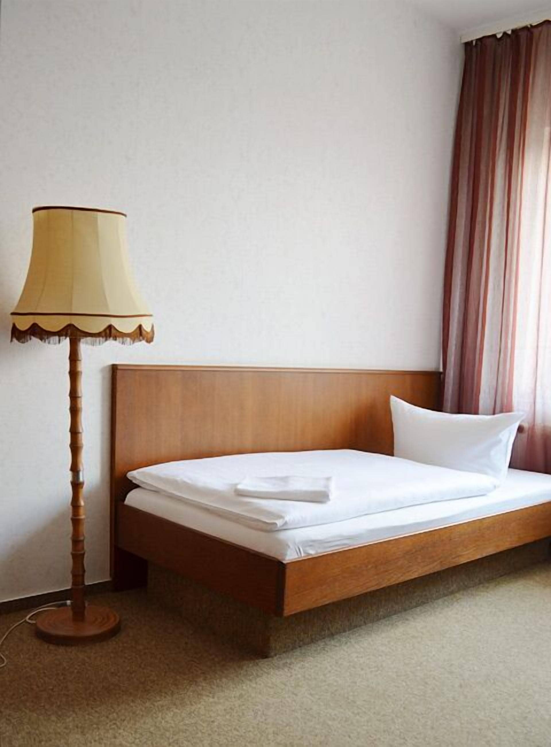 Una cama bien hecha con sábanas blancas y una toalla doblada, un cabecero de madera, una lámpara de pie con pantalla color beige y una cortina a rayas.