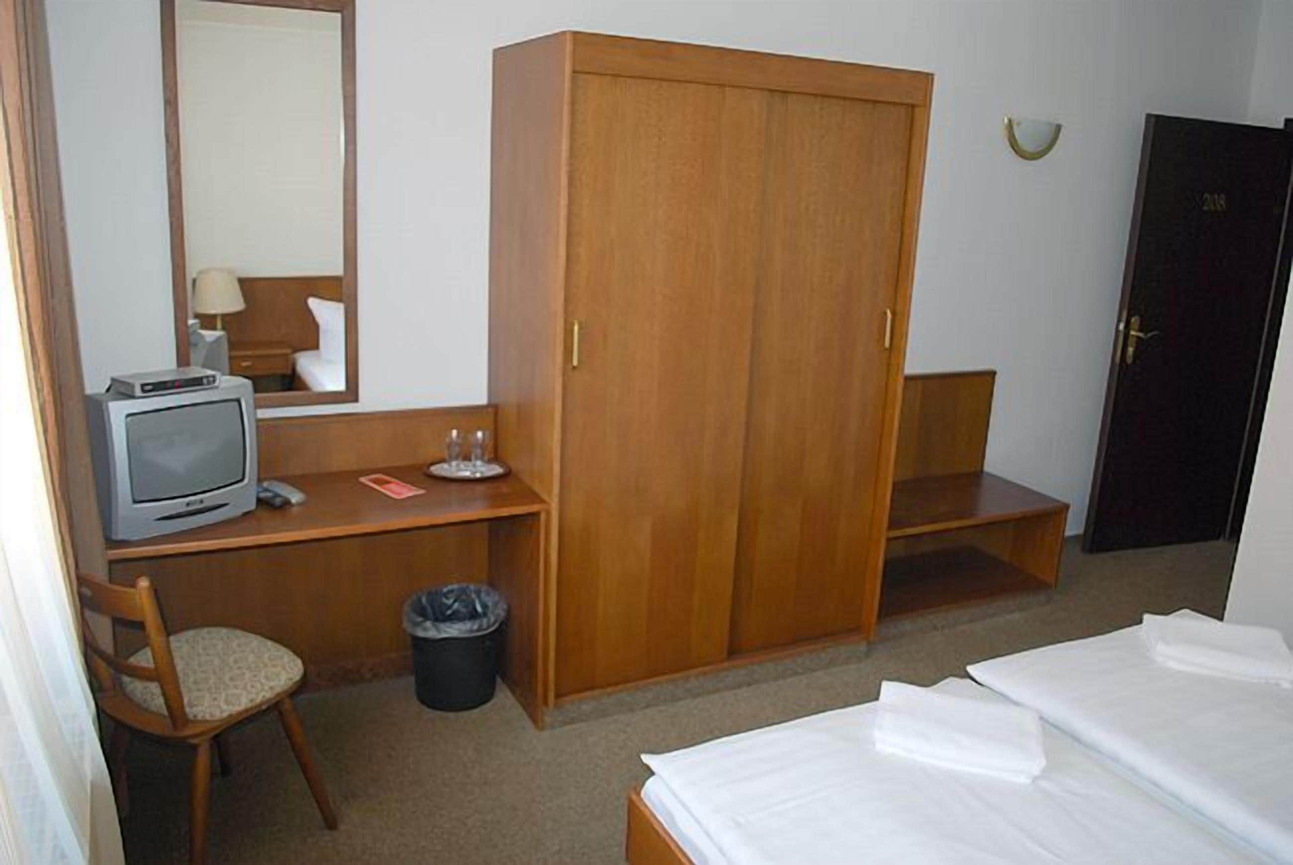 Quarto de hotel com uma escrivaninha de madeira, televisão, guarda-roupa, cadeira e cama.