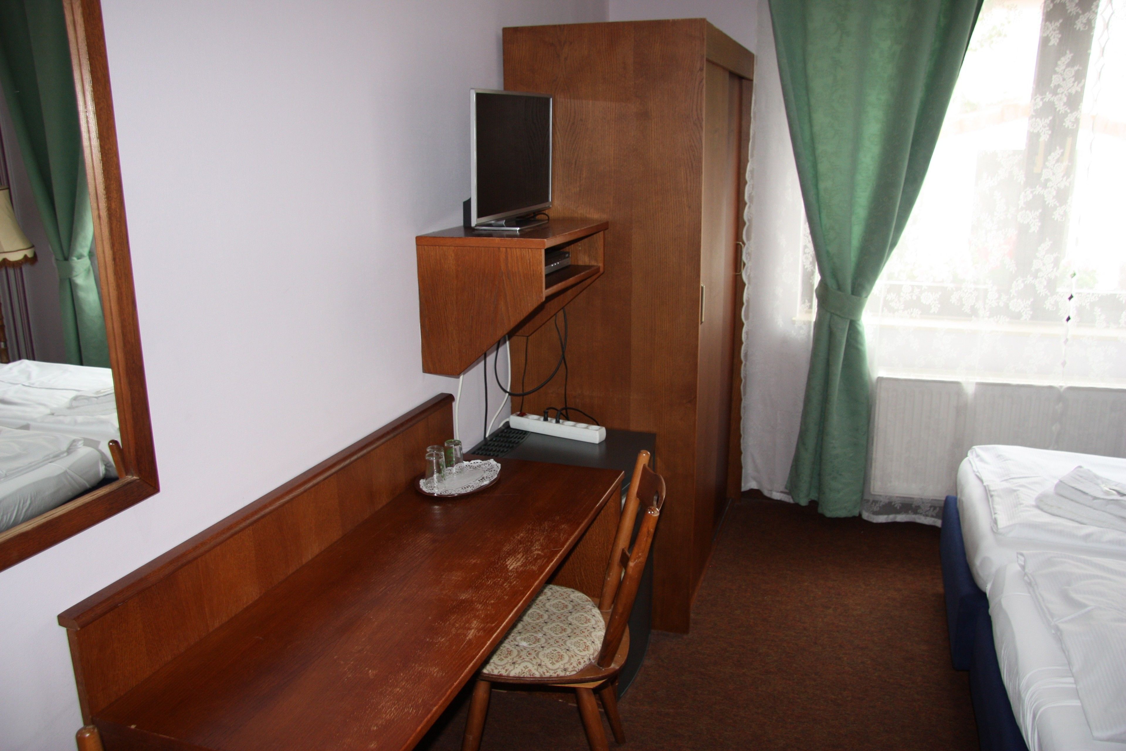 A hotel room with a wooden desk, a television on a shelf, a bed, and a window with curtains.