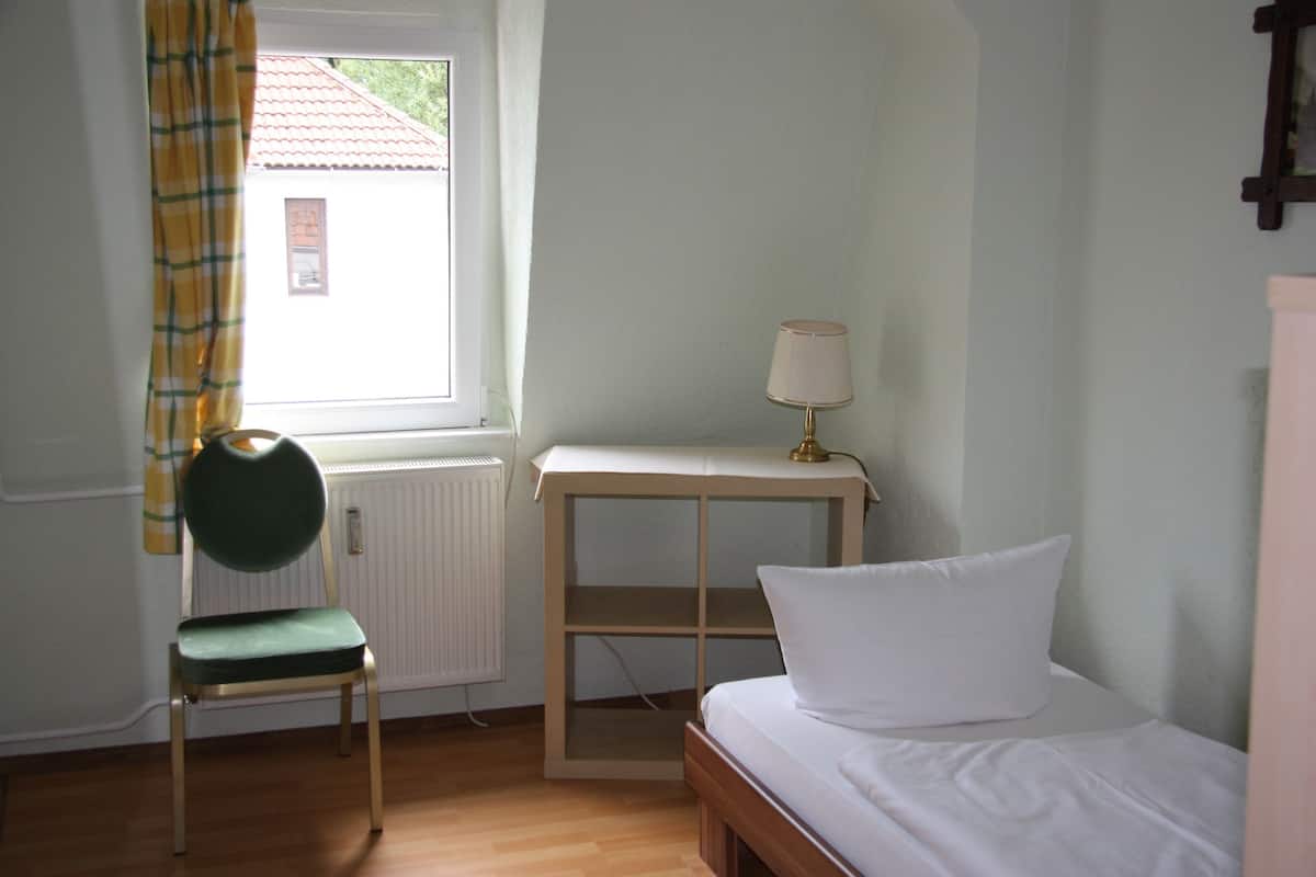 A room with a bed, a chair, a table with a lamp, and a window with curtains.