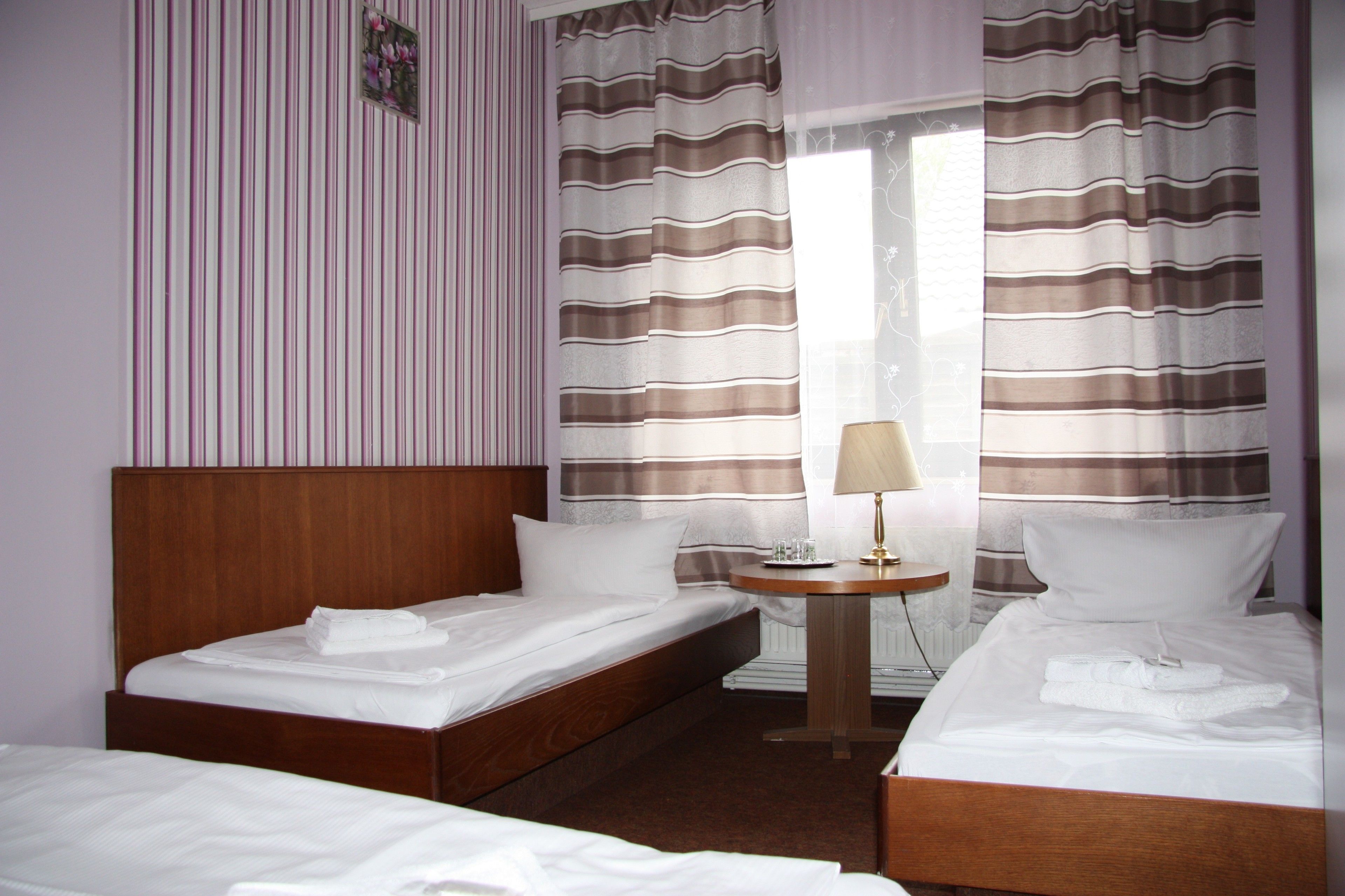 A hotel room with two single beds, a wooden headboard, a small bedside table with a lamp, and striped curtains.