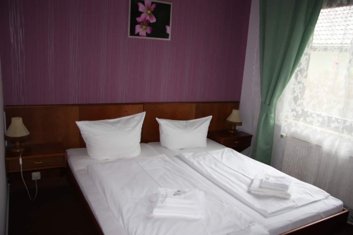 A double bed with white linens, two bedside tables with lamps, a floral picture on the wall, and a window with sheer curtains.