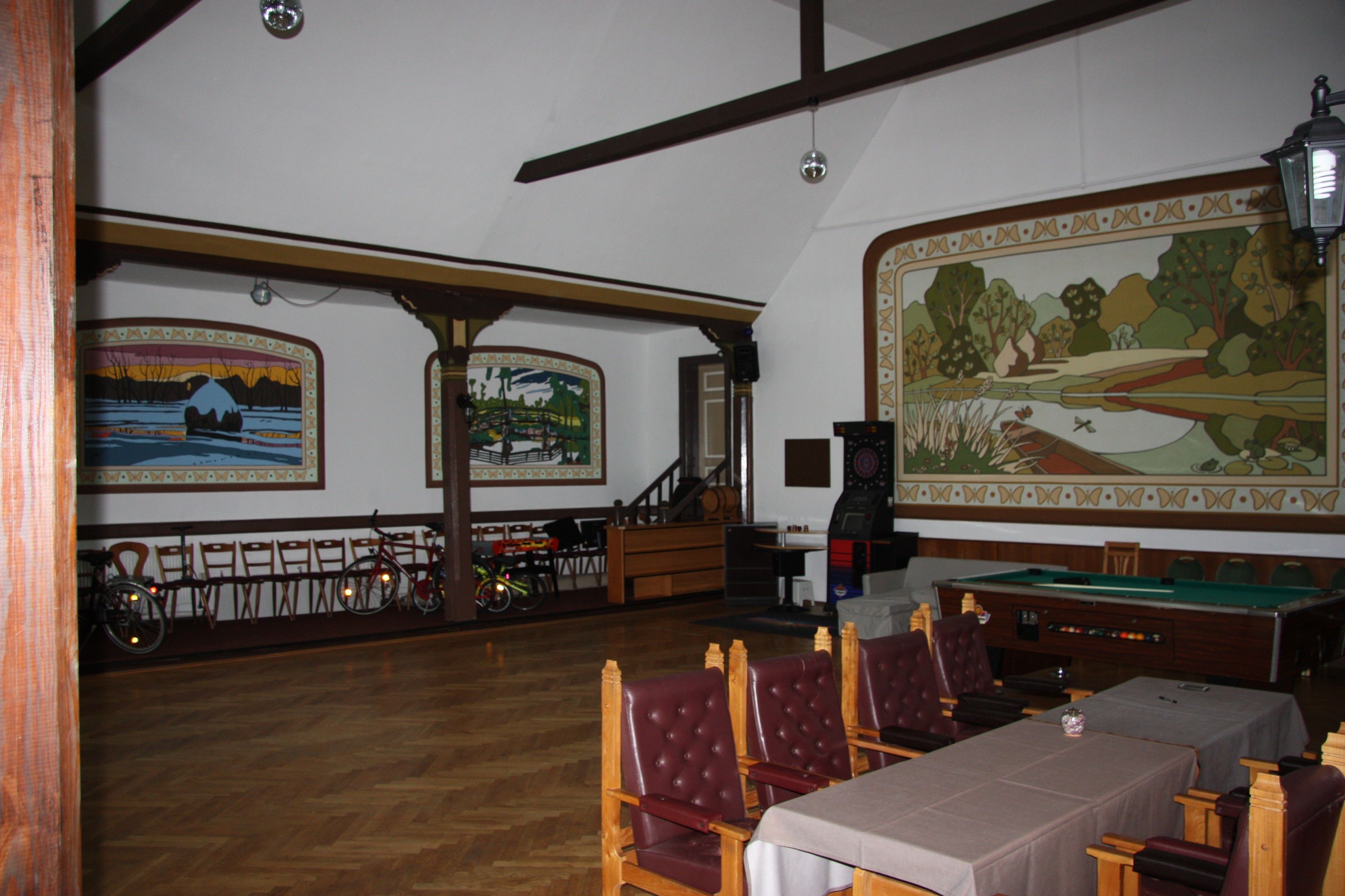 Une salle de billard avec des tables et des chaises en bois, une table de billard et deux grandes peintures de paysages accrochées aux murs.