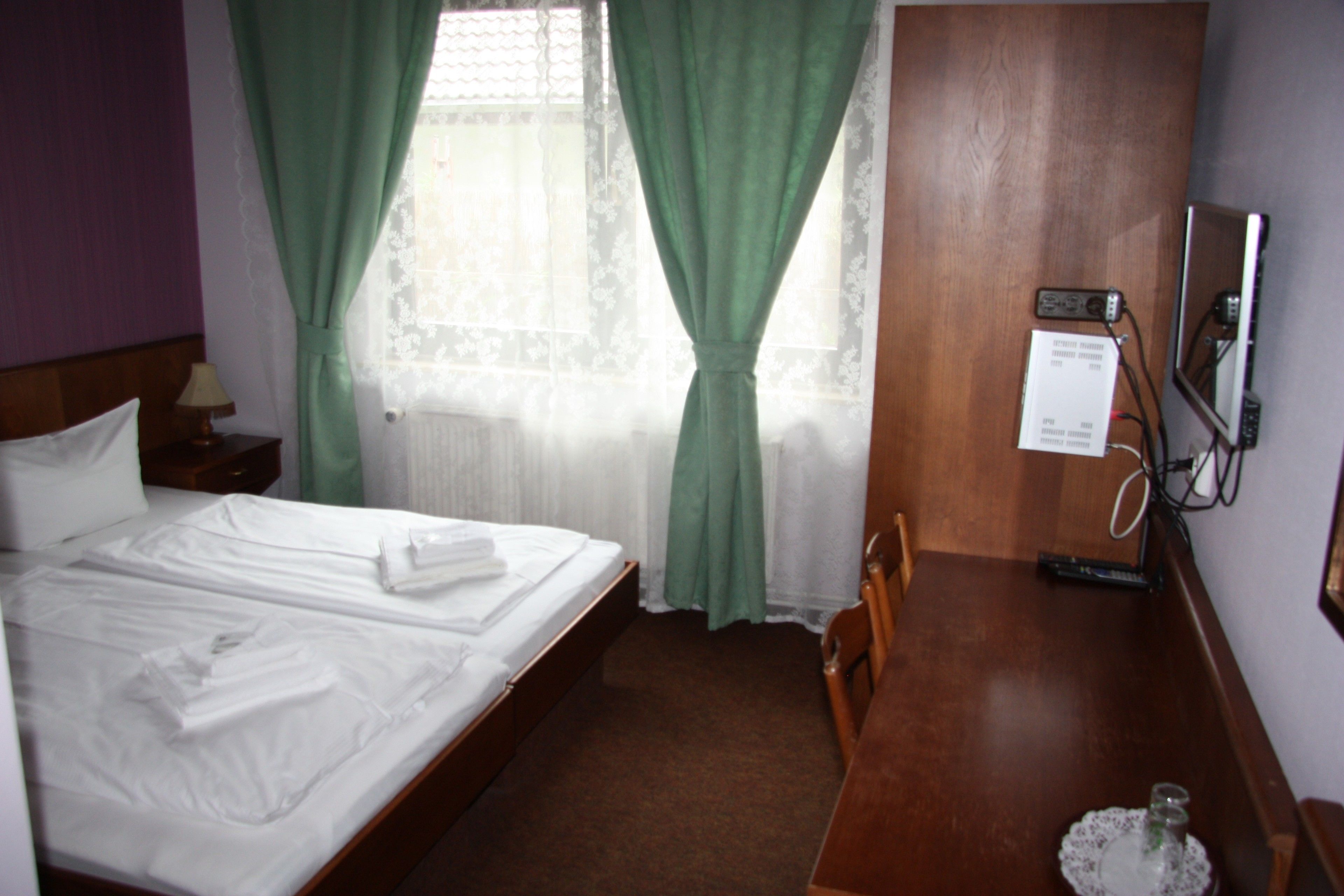 A hotel room with a neatly made bed, a wooden desk, a chair, a lamp, and a window with curtains.