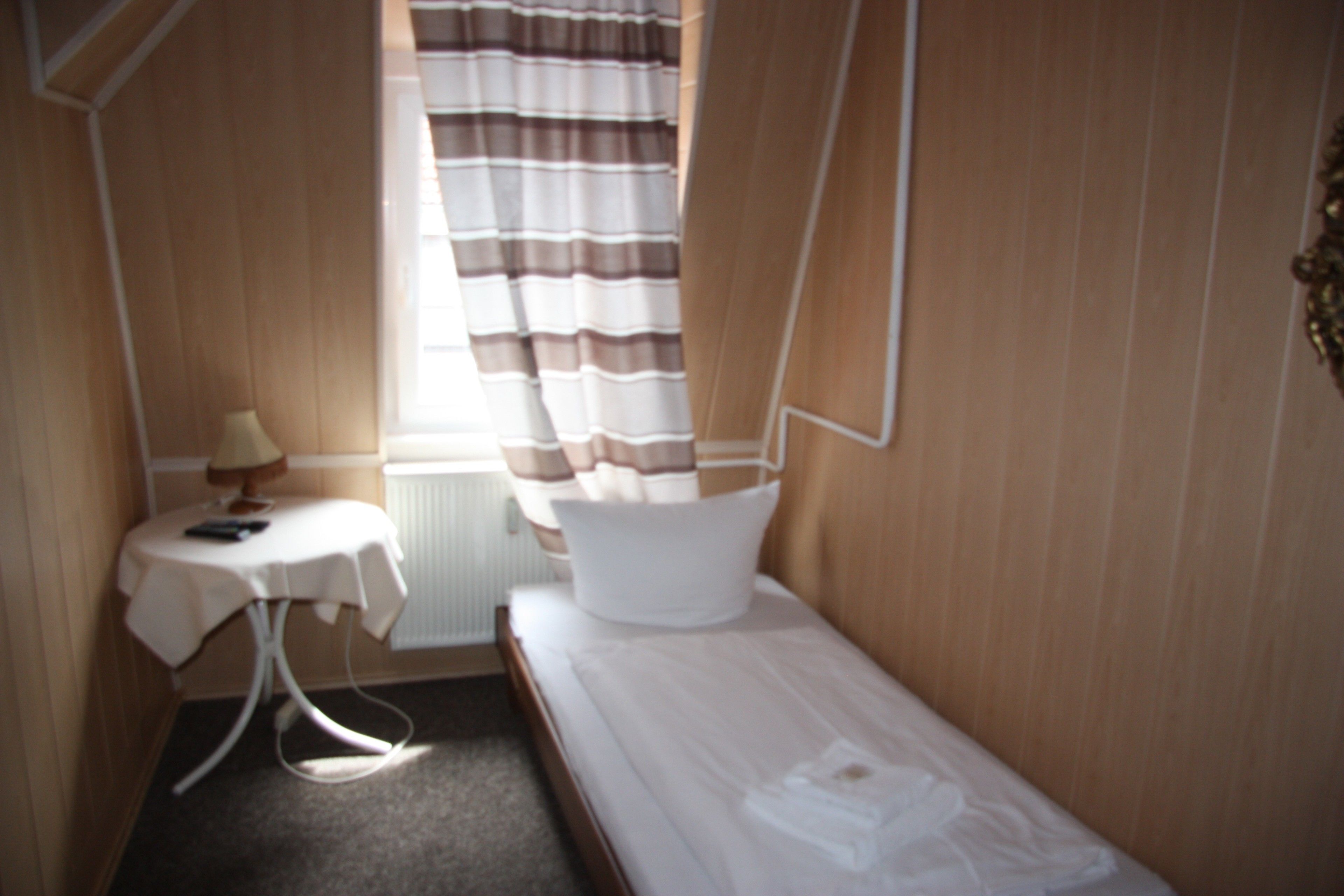 A small, neatly arranged room with a single bed, a small table with a lamp, and a window with curtains.