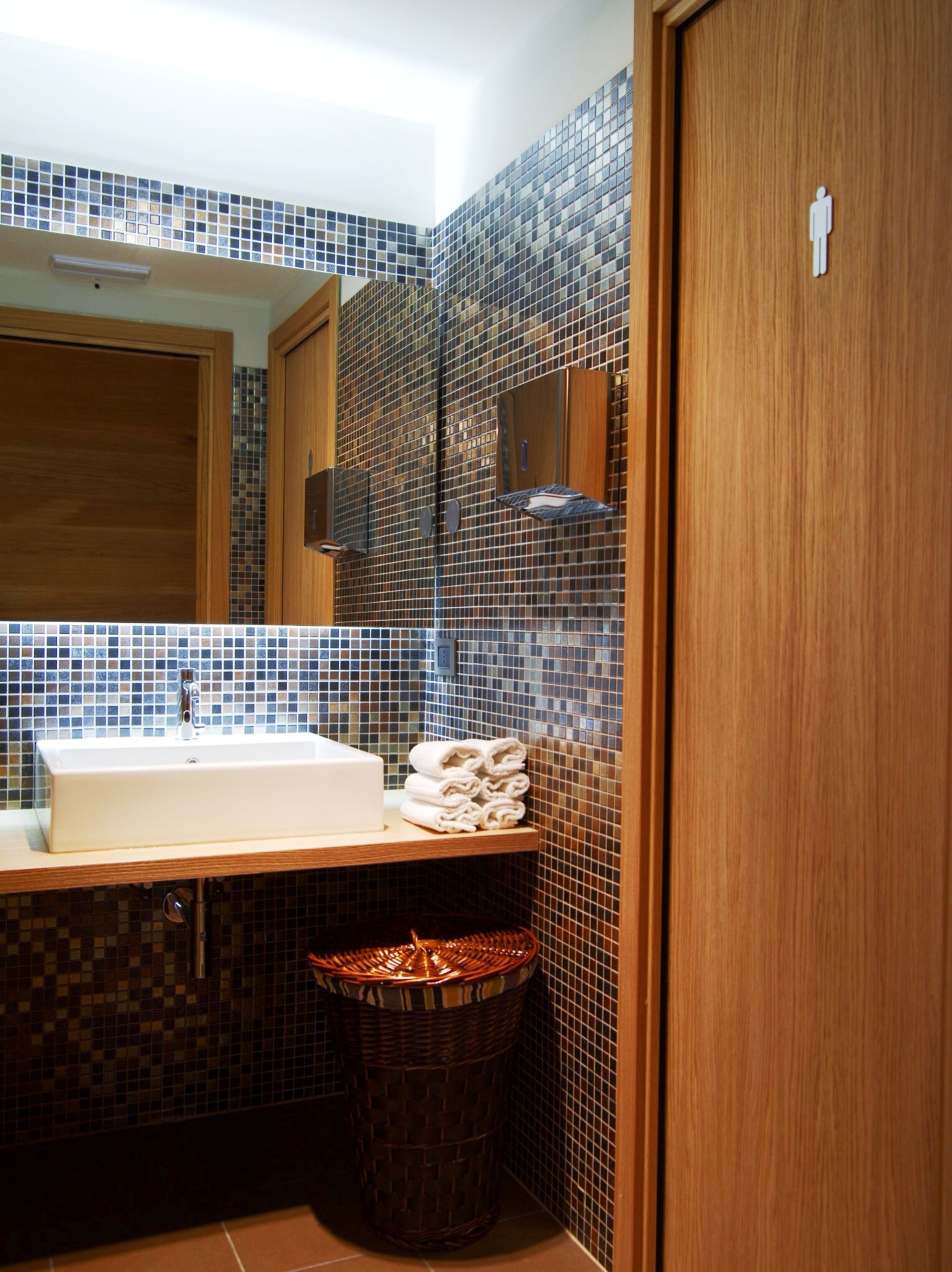 Une salle de bain avec un mur recouvert de carreaux derrière le lavabo, un lavabo, un miroir, un porte-serviettes et une poubelle.