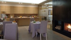 A modern dining area with a kitchen in the background, a fireplace, and a buffet with various food items. - Hotel Natura (Castel di Sangro)