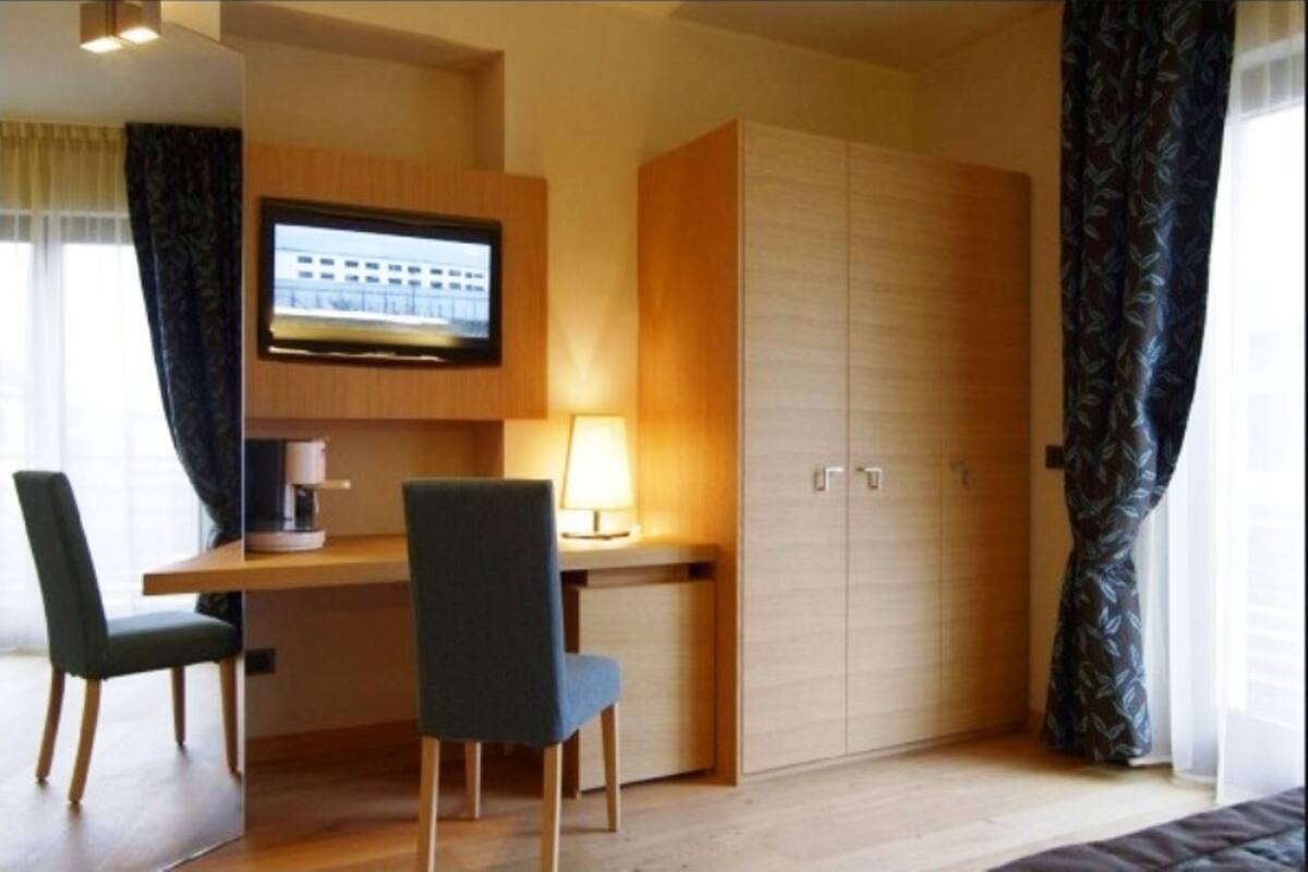 A hotel room with a wooden desk, two chairs, a television mounted on the wall, and a wardrobe.