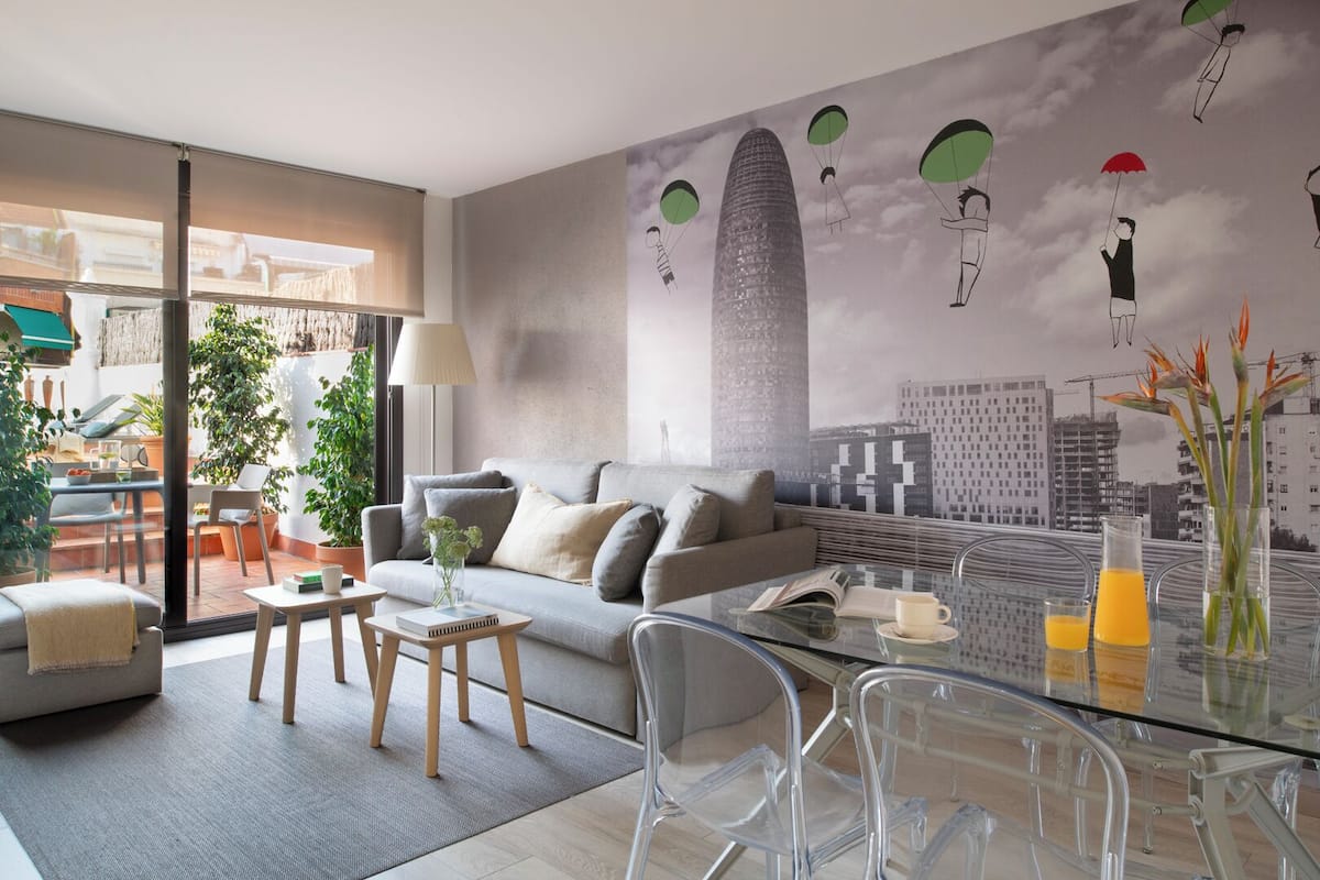 A modern living room with a sofa, coffee table, and chairs. There is a glass table with drinks and a vase with flowers. A mural of parachuting figures and buildings is on the wall.