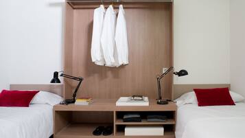 A modern hotel room with two beds, a wooden wardrobe, and a desk with a lamp.