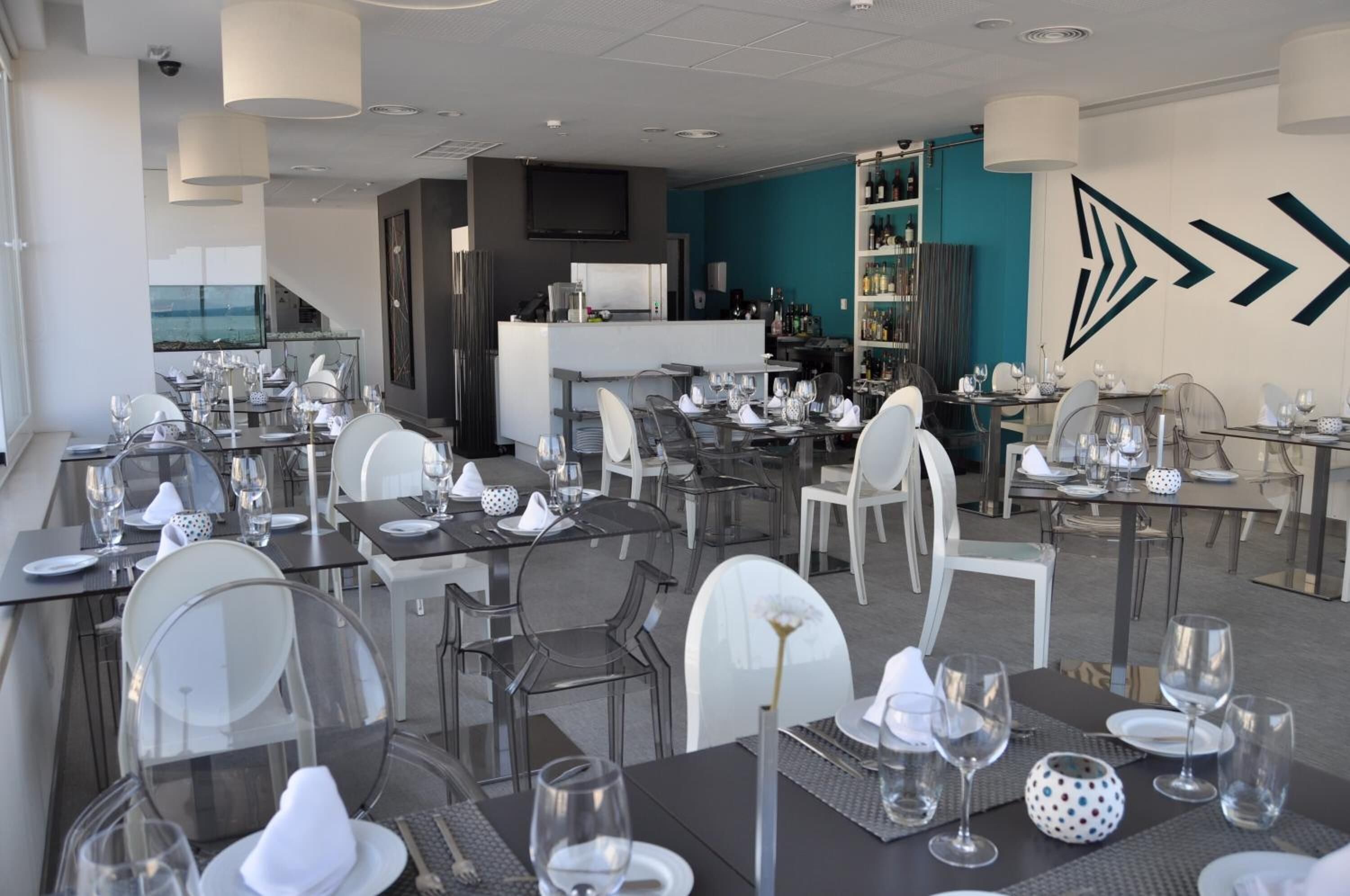 Un restaurant moderne avec des tables et des chaises en verre, un coin bar et un mur d’accent de couleur turquoise.