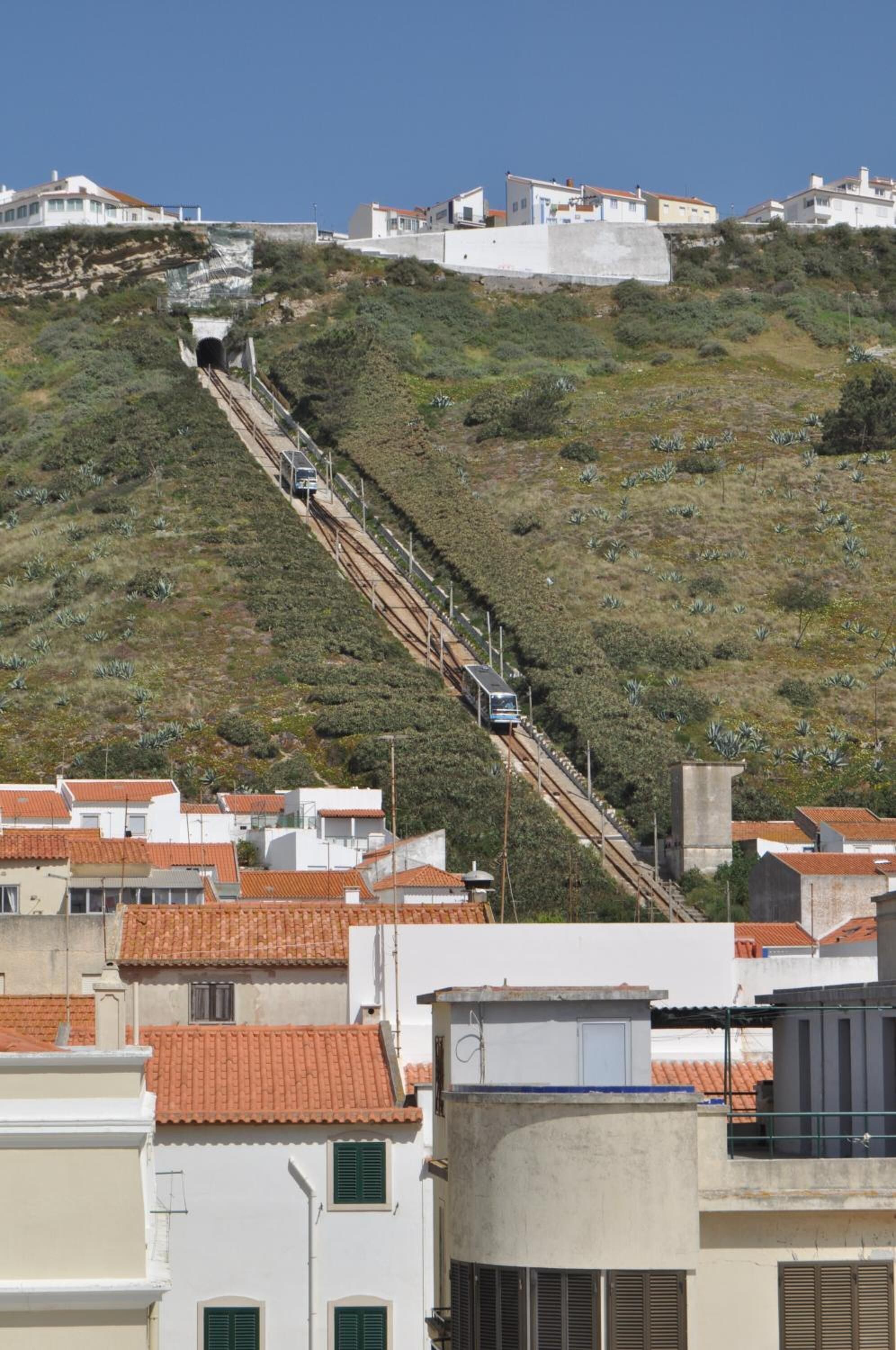 A hillside with a railway track ascending towards a tunnel entrance, surrounded by residential buildings with red-tiled roofs.