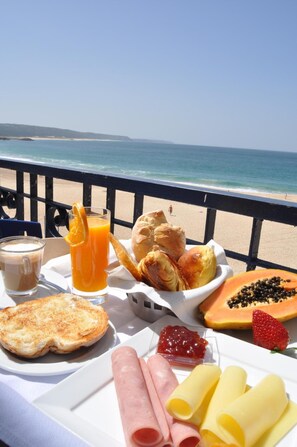 A breakfast table with croissants, a glass of orange juice, a slice of toast, a plate of ham, cheese, and a fruit salad on a balcony overlooking the beach. - Hotel Mar Bravo (Nazaré)
