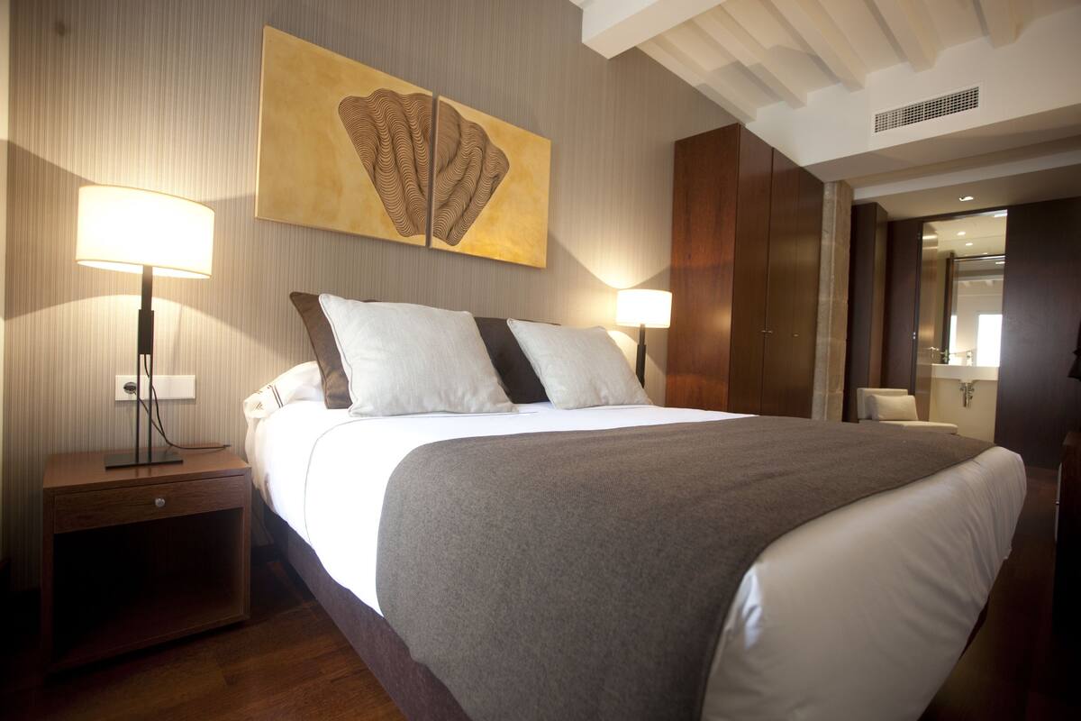 A hotel room with a large bed, two bedside lamps, a nightstand, a wall-mounted artwork, and a wooden wardrobe.