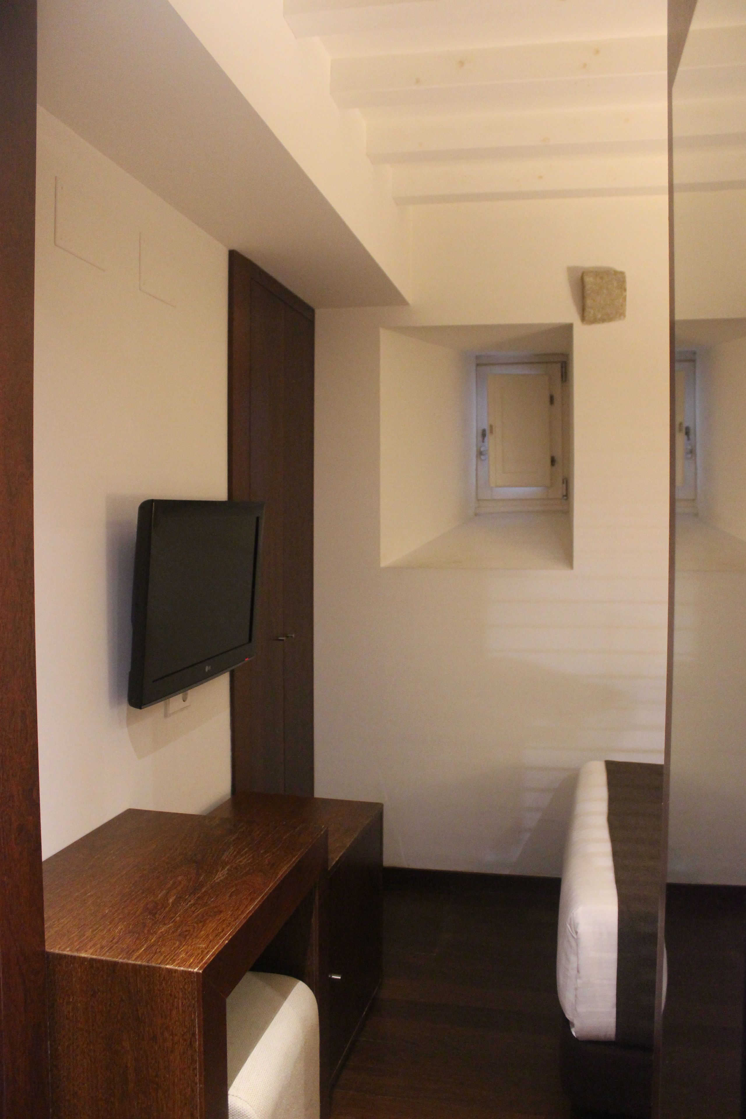 Una habitación moderna con una televisión de pantalla plana montada en un mueble de madera, una ventana pequeña y una cama con colchón blanco.