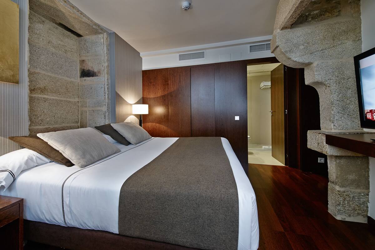 A bedroom with a bed, a nightstand, a lamp, a stone wall, a wooden door, and a television.