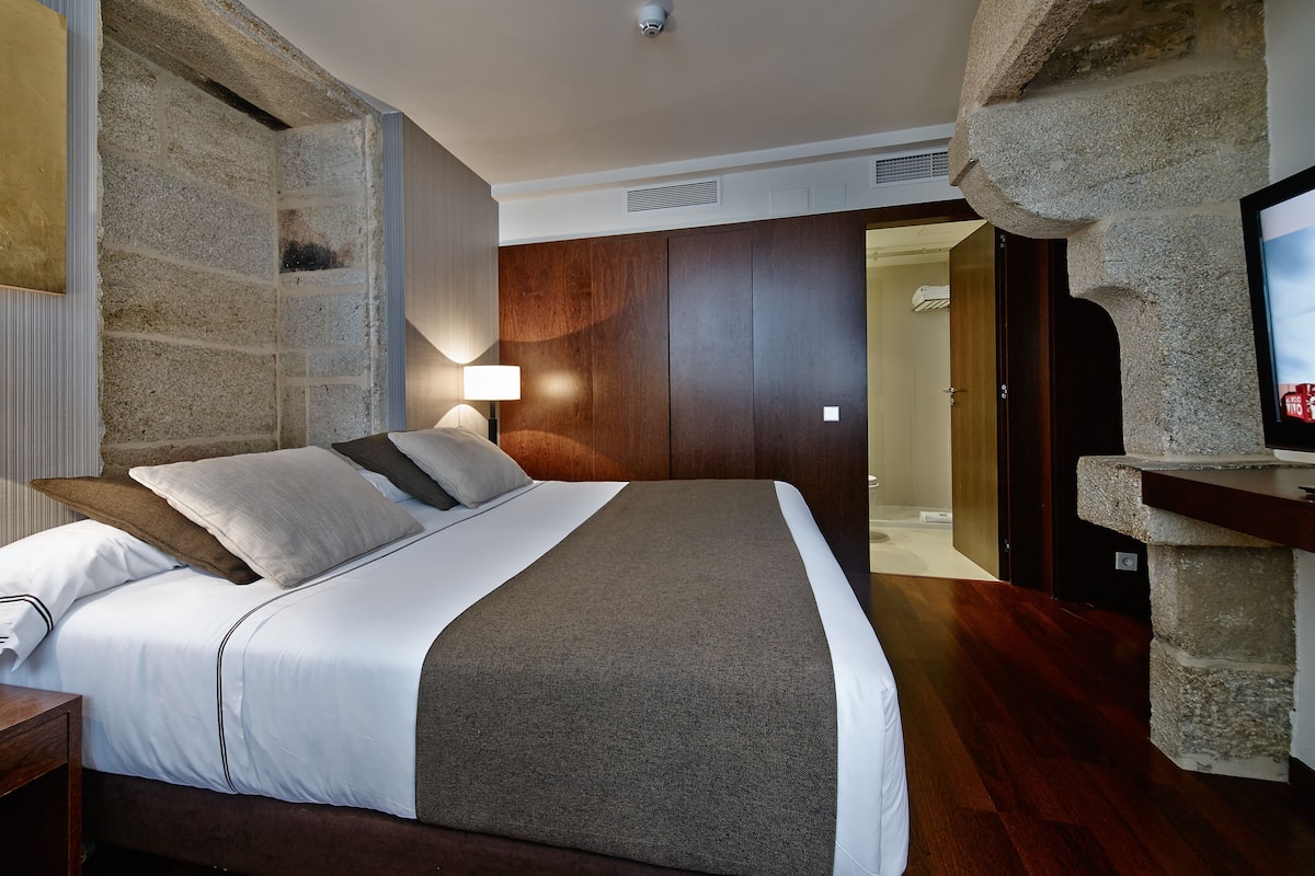 A bedroom with a bed, a nightstand, a lamp, a stone wall, a wooden door, and a television.