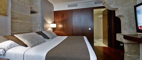 A modern hotel room with a large bed, a stone accent wall, a wooden wardrobe, a bedside lamp, and a television on a stone shelf.