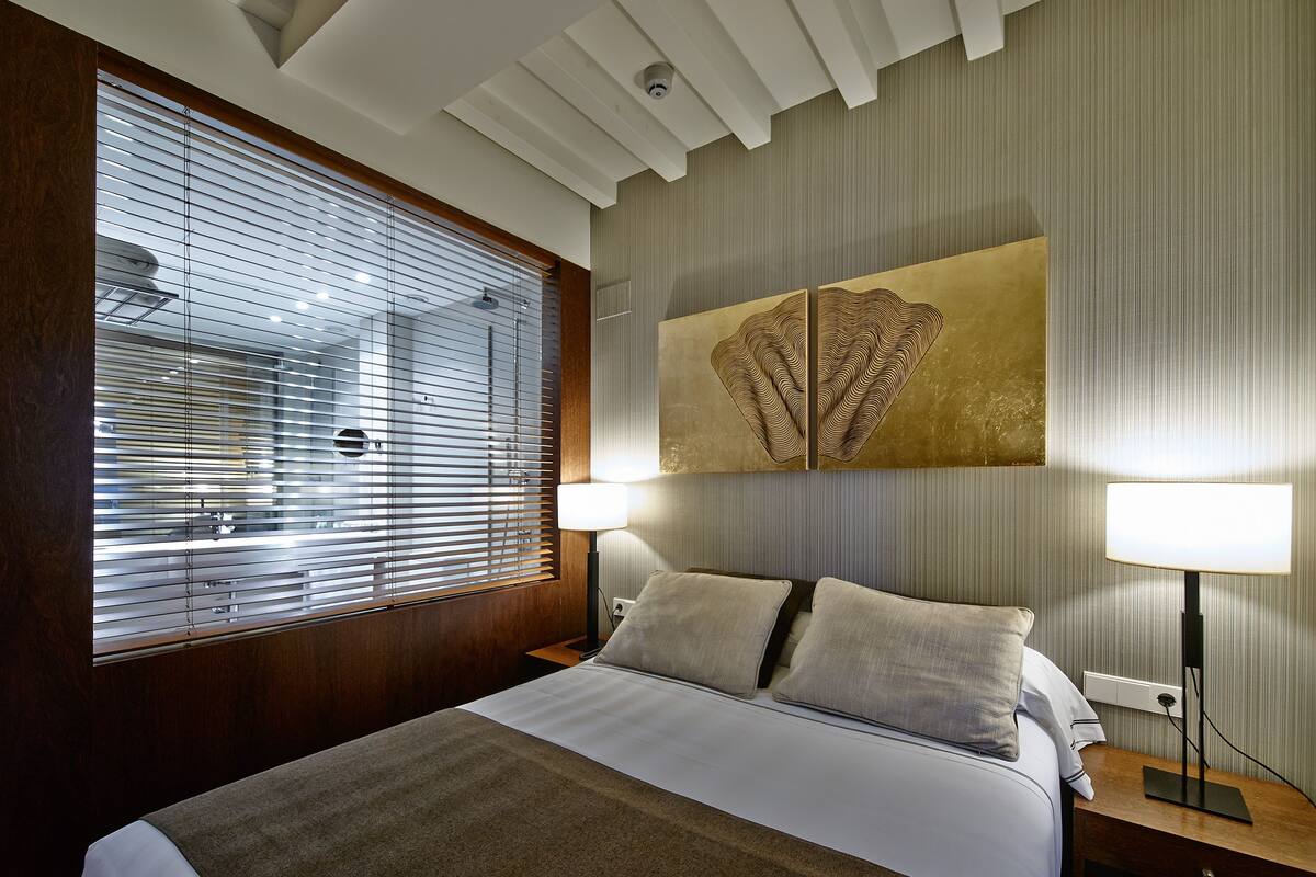 A hotel room with a large bed, two bedside lamps, a window with blinds, and a wall-mounted artwork.