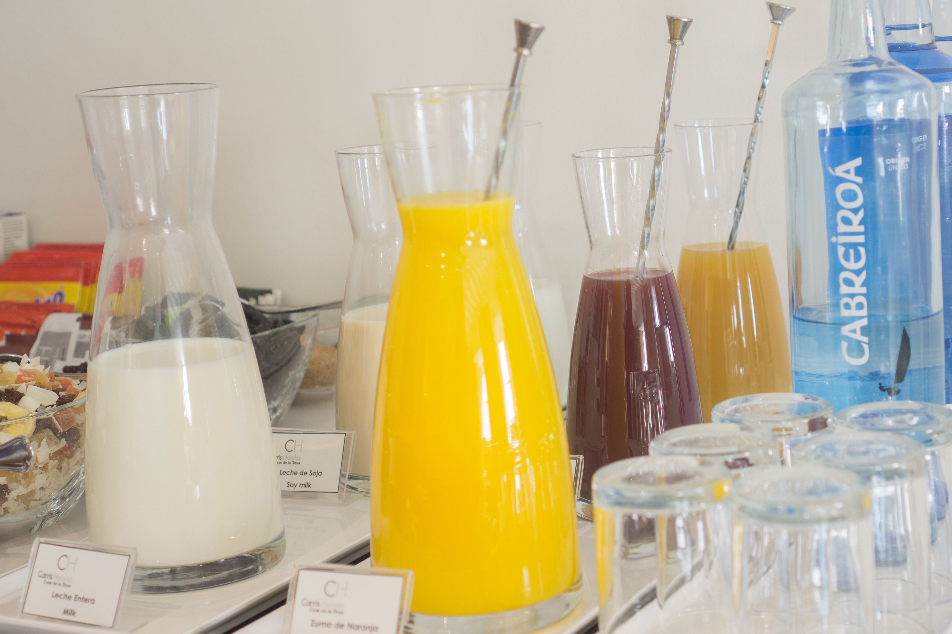 Variedad de bebidas en diferentes recipientes, como leche, zumo de naranja y otras opciones, expuestas sobre una mesa con etiquetas.