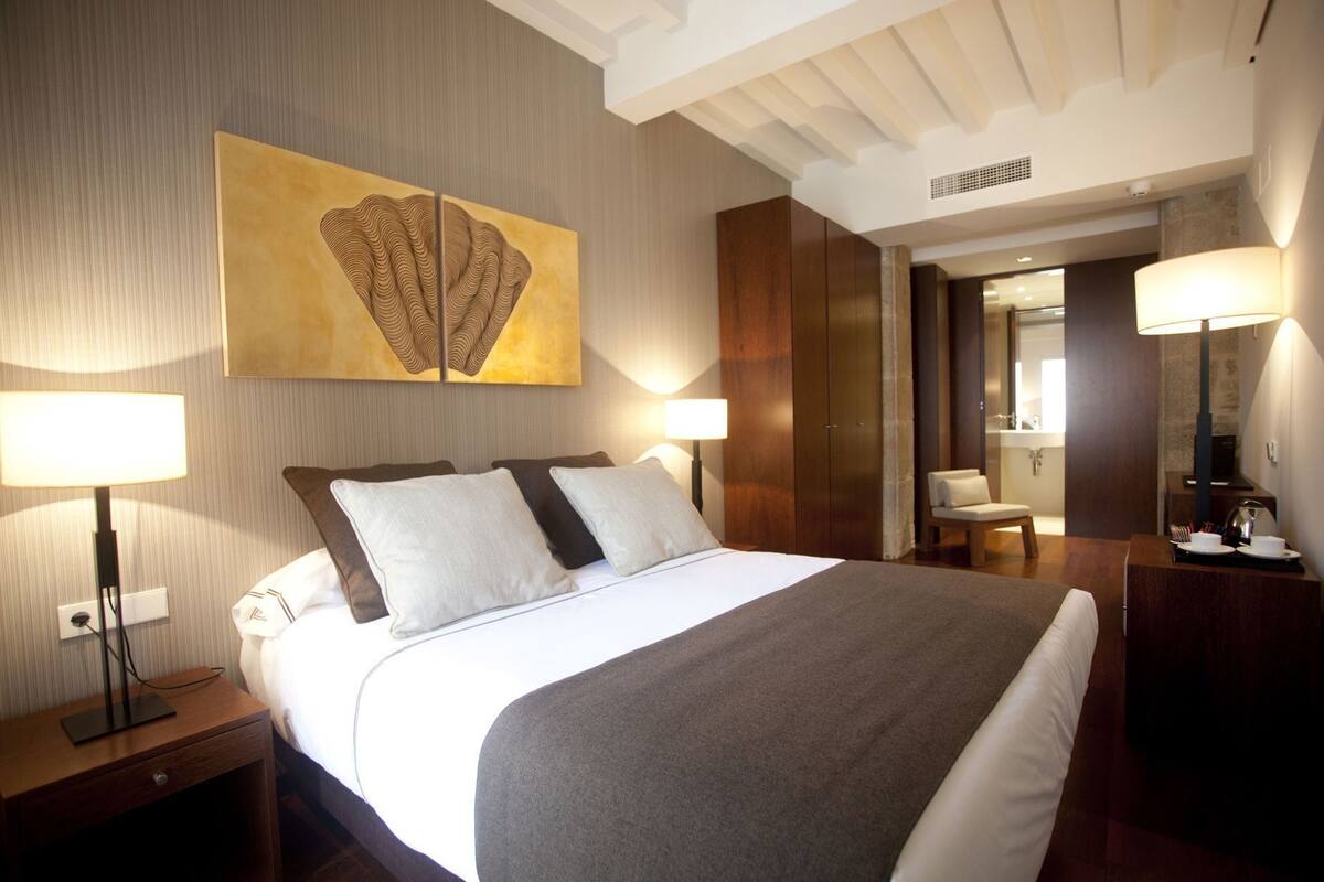 A hotel room with a large bed, two bedside lamps, a wall-mounted artwork, a mirror, and a wooden nightstand.