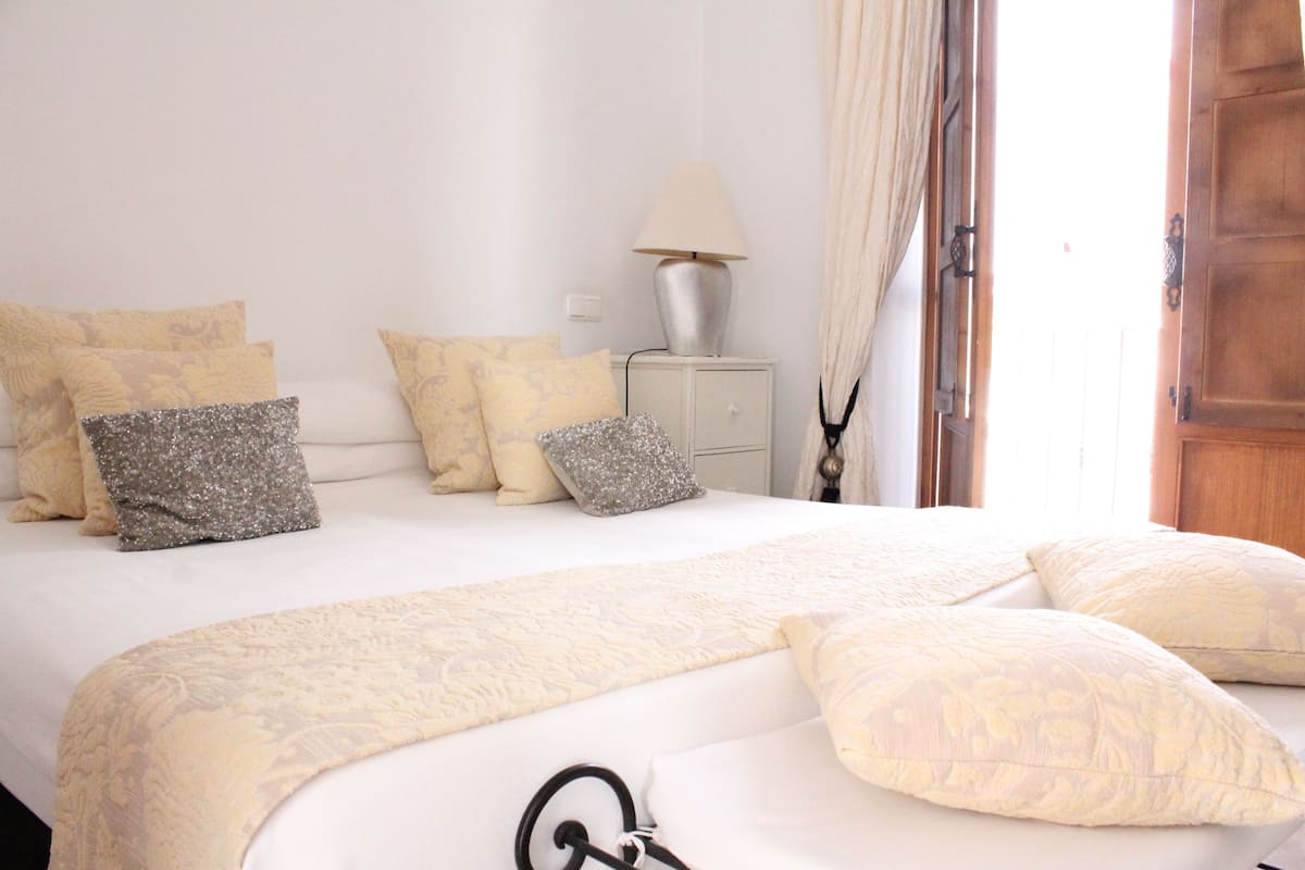 Een net opgemaakt bed met een bedspreidek en meerdere kussens, een nachtkastje met een lamp, en een houten deur op de achtergrond.
