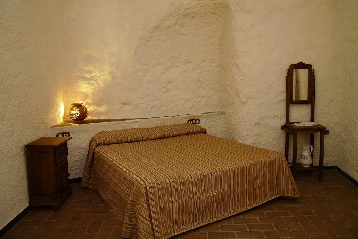 Ein Zimmer mit einem Bett, einem Nachttisch, einer Lampe, einem Spiegel und einem kleinen Schrank.