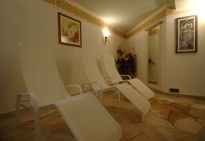 Een kamer met vier witte loungestoelen op een rij, een wandlamp en twee florale schilderijen in lijsten.