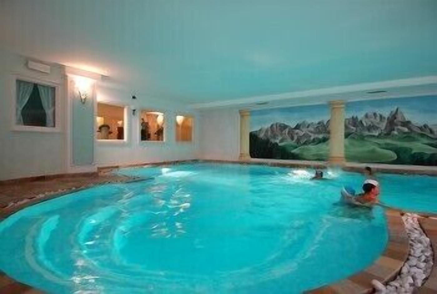 An indoor swimming pool with a mountain landscape mural on the wall, a person swimming, and a stone border around the pool.