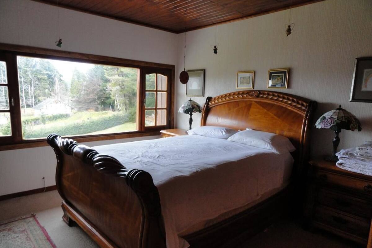 Une chambre avec un grand lit, une fenêtre donnant sur un paysage verdoyant, une tête de lit en bois et une table de chevet avec une lampe.