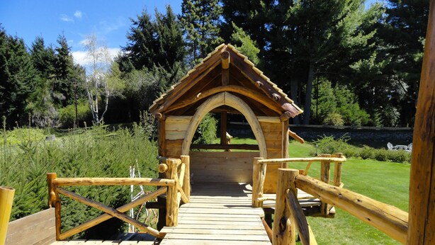A wooden bridge leading to a rustic wooden structure with a pointed roof, surrounded by greenery.