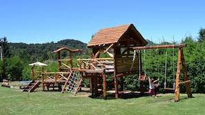 A wooden play structure with a climbing wall and a slide, set in a grassy field with trees in the background.