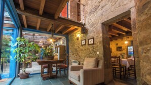 Un interior acogedor con una pared de piedra, vigas de madera en el techo, una zona de comedor con una mesa y sillas, y una planta en maceta.