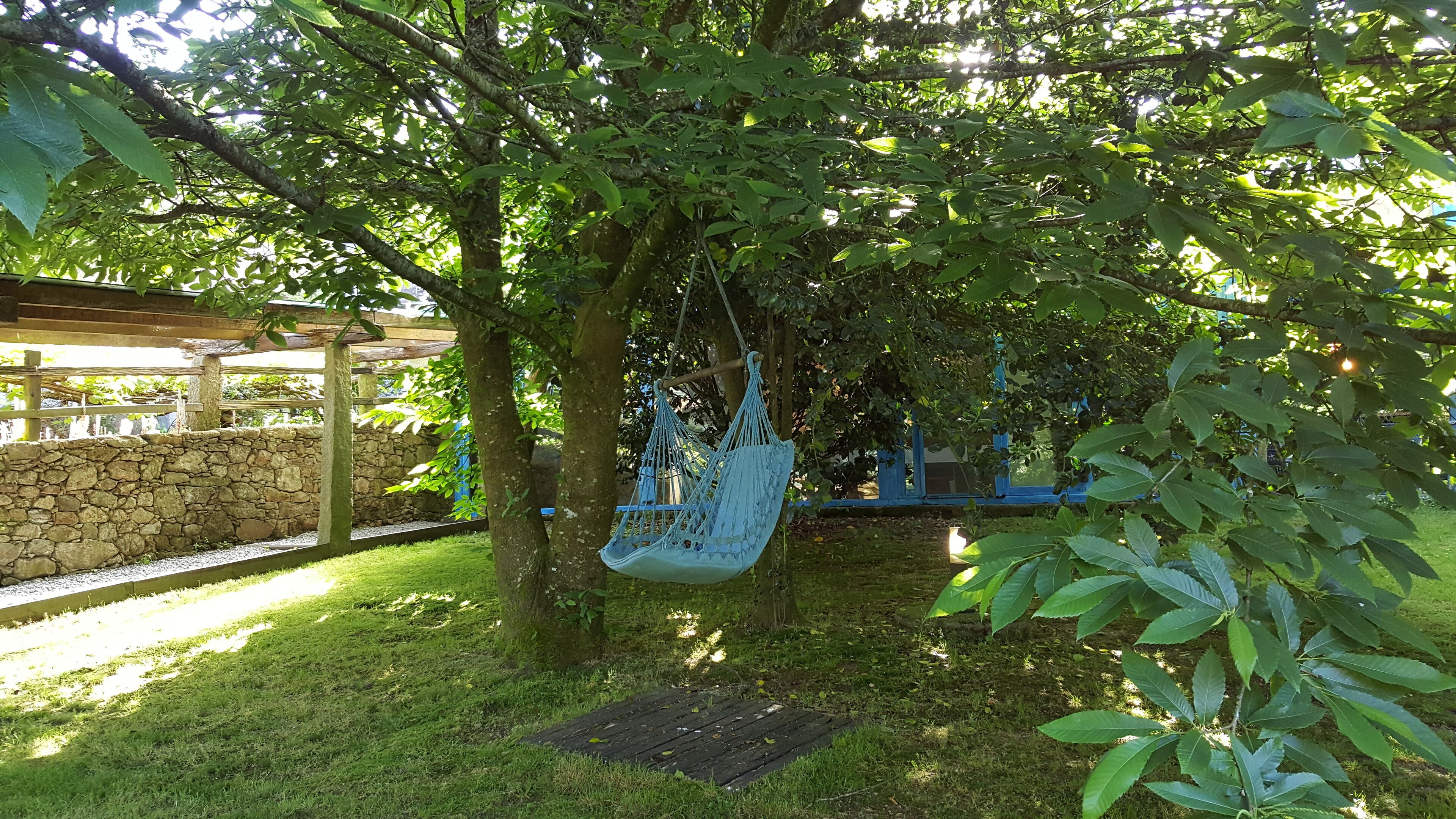 A hammock hanging from a tree in a garden area with a stone wall and a wooden pergola in the background.