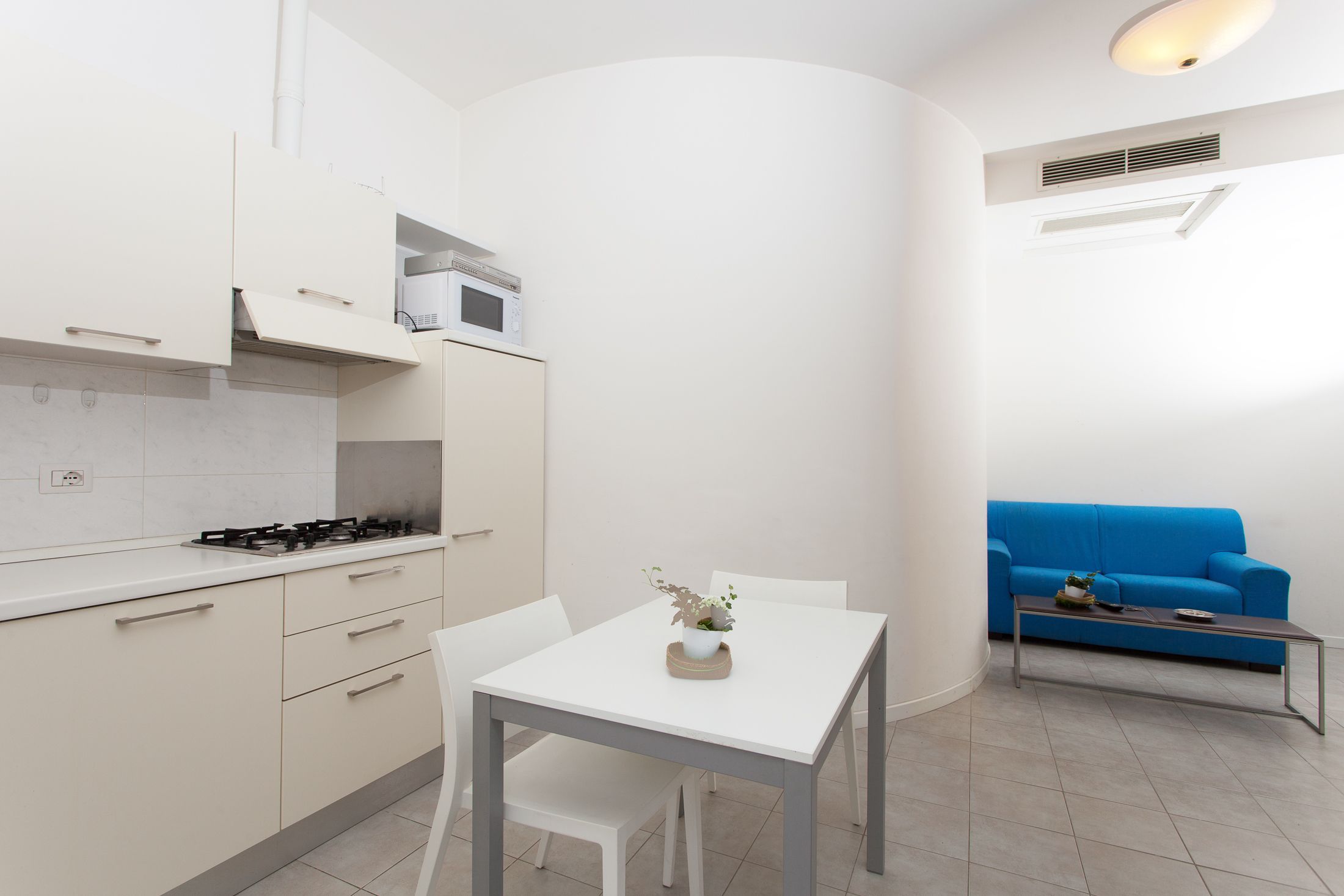 Une cuisine moderne avec des meubles blancs, une cuisinière à gaz et un four à micro-ondes. On y trouve une table à manger avec des chaises, ainsi qu’un canapé bleu dans l’espace salon.