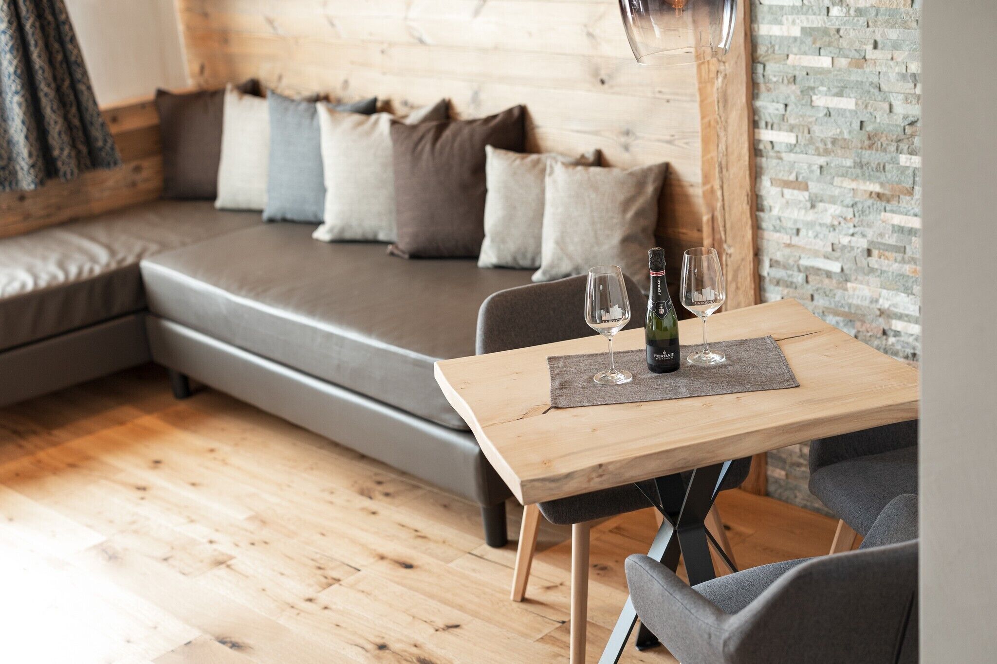 A cozy room with a wooden table set for two, a grey sofa with cushions, and a stone wall.
