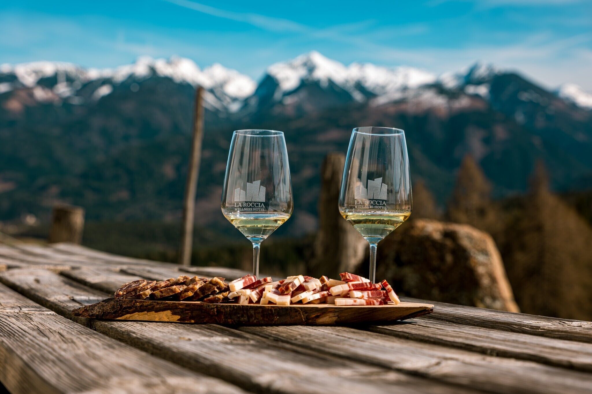 Deux verres de vin blanc sur une table en bois, accompagnés d’une assiette de charcuterie, le tout sur fond de paysage montagneux.