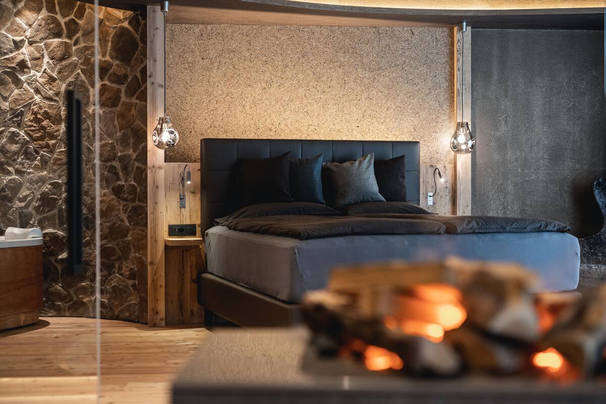 A modern bedroom with a stone wall, a wooden headboard, and a bed with dark bedding.