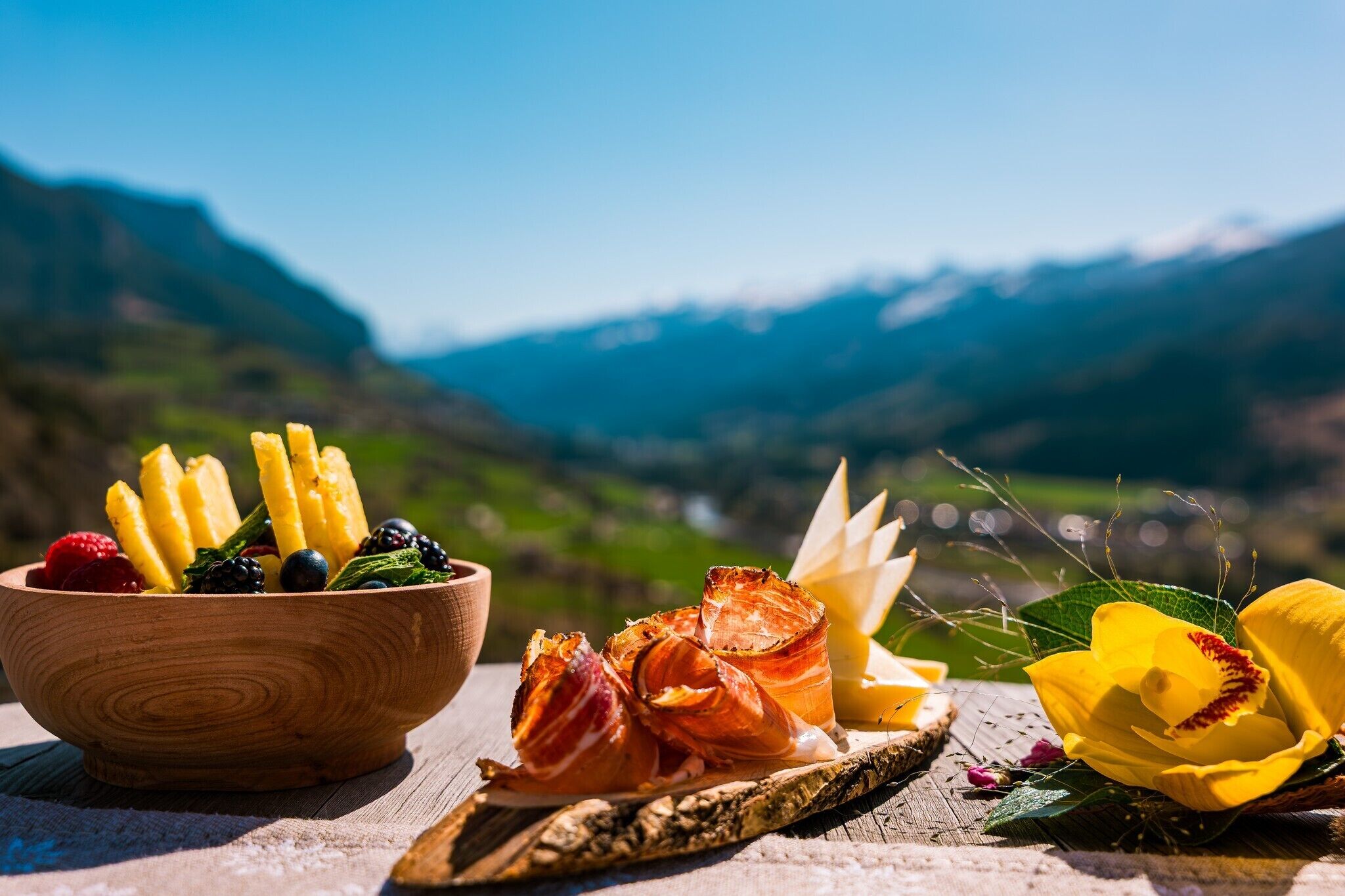 A wooden bowl filled with fruits and vegetables, a wooden cutting board with a flower, and a wooden spoon on a table with a mountainous backdrop.