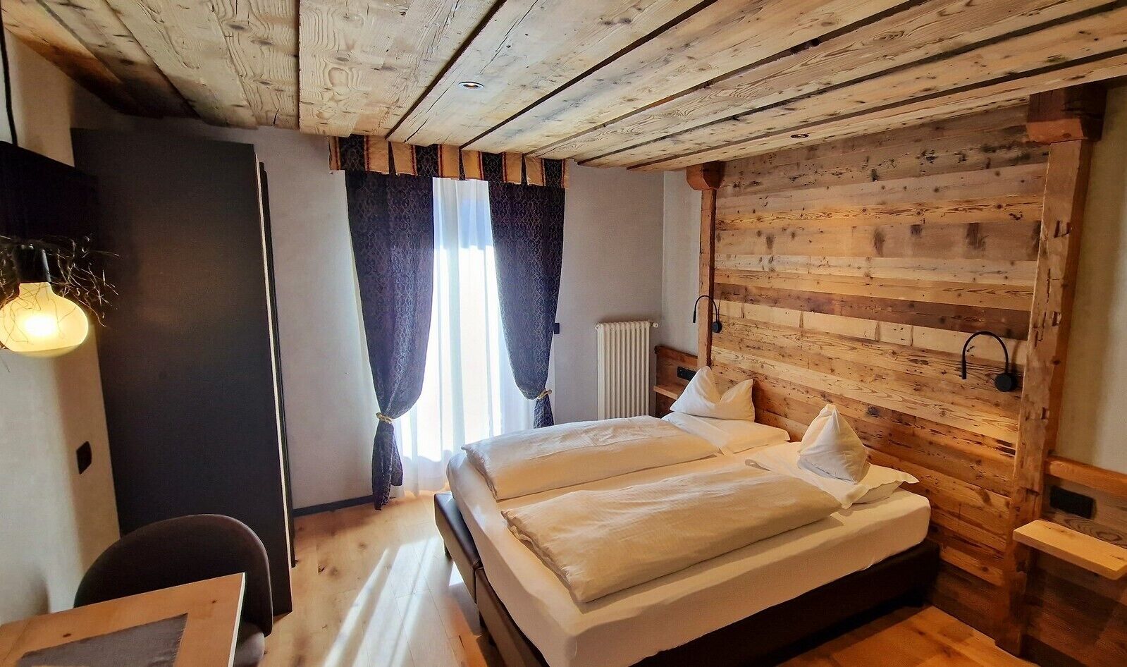 A cozy room with a wooden ceiling, a bed with white linens, a desk with a chair, and a window with curtains.