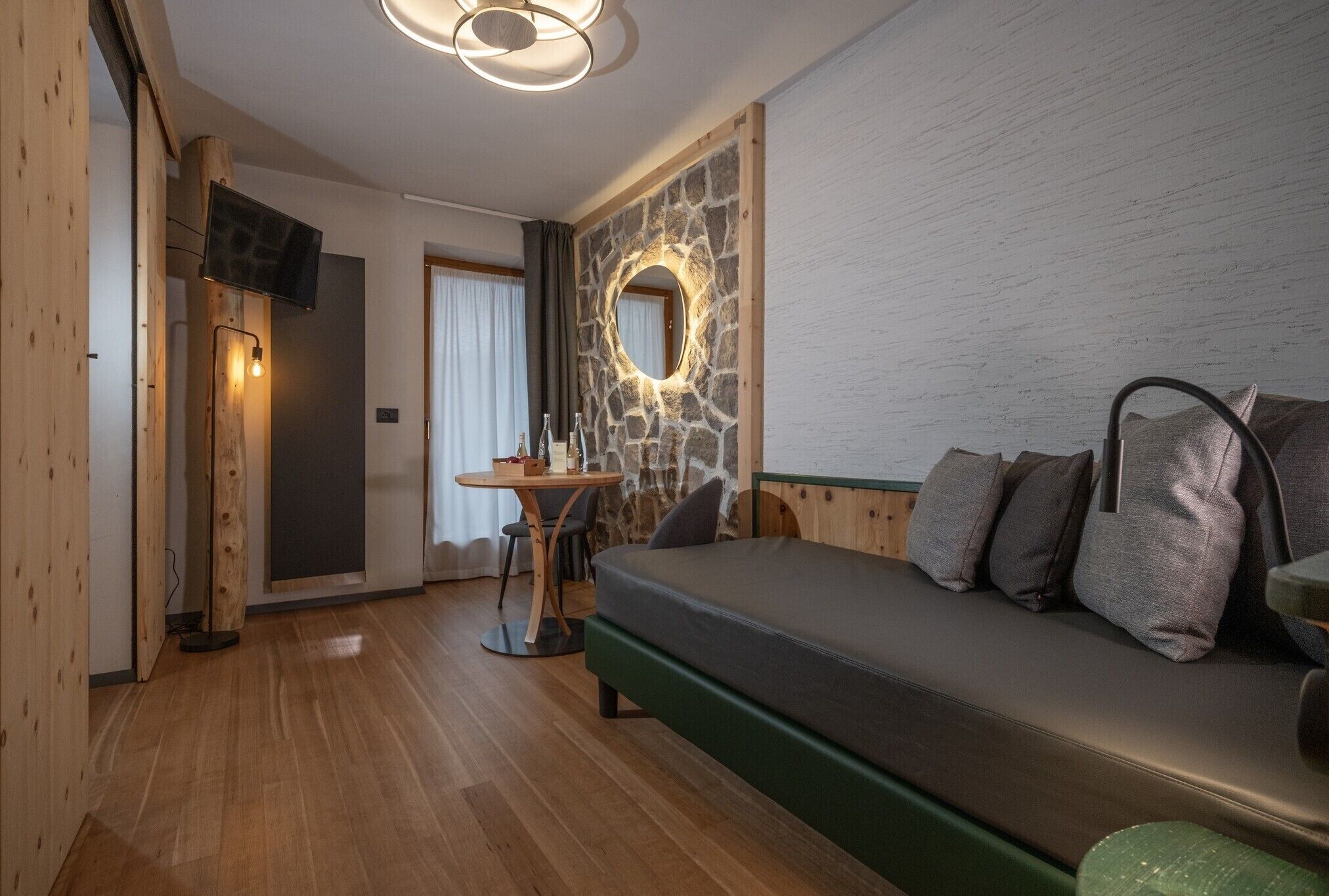 Una habitación acogedora con suelo de madera, un sofá verde con cojines, una pequeña mesa redonda, una pared de piedra con un espejo circular y un televisor sobre un mueble de madera.