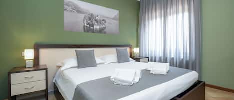 A neatly made bed with white linens and gray pillows, a bedside table with a lamp, and a large window with curtains.