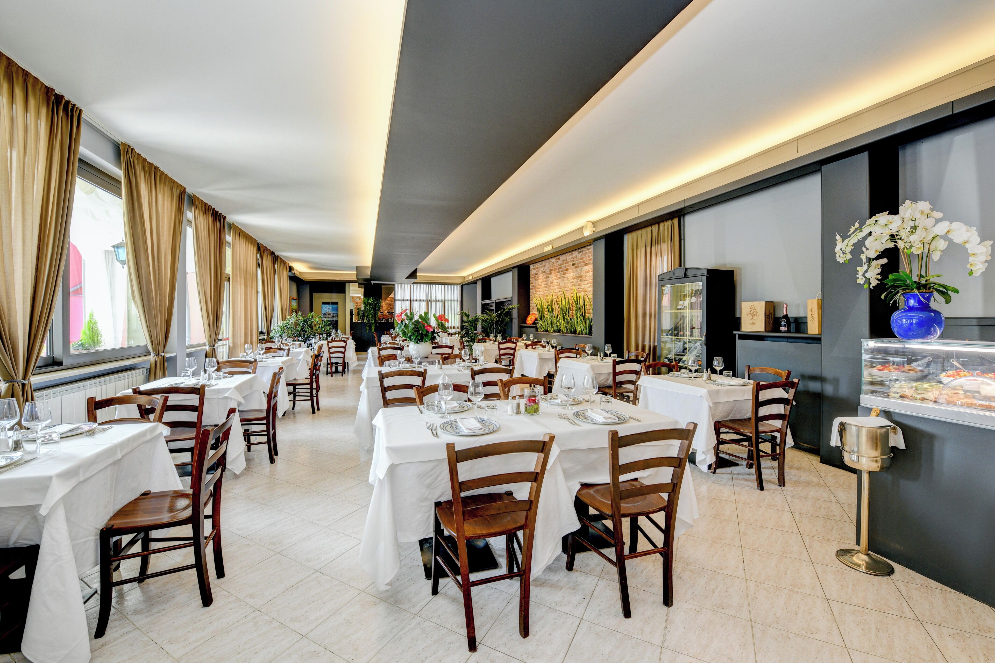 Un restaurante espacioso con varias mesas y sillas, una vitrina de postres y grandes ventanales con cortinas.