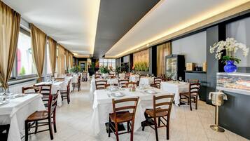 Un restaurante espacioso con varias mesas y sillas, una vitrina de postres y grandes ventanales con cortinas.