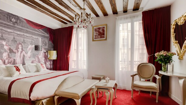 Una habitación de hotel con una cama grande, una mesita pequeña, una silla, un escritorio, un espejo y una araña.